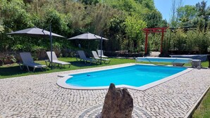 A swimming pool with lounge chairs and umbrellas, surrounded by greenery and a stone pathway.