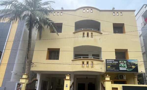 Un edificio de varios pisos con un balcón en el segundo nivel, un letrero que anuncia 'Miracle Home Stays' con información de contacto, y una palmera en primer plano.
