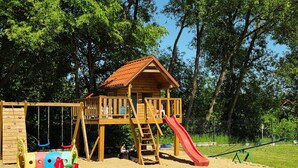 Une structure de jeu en bois, dotée d’un toboggan et d’une zone d’escalade, entourée de sable et d’arbres.