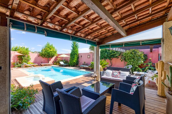 An outdoor pool area with lounge chairs and a table, surrounded by greenery and a wooden deck.
