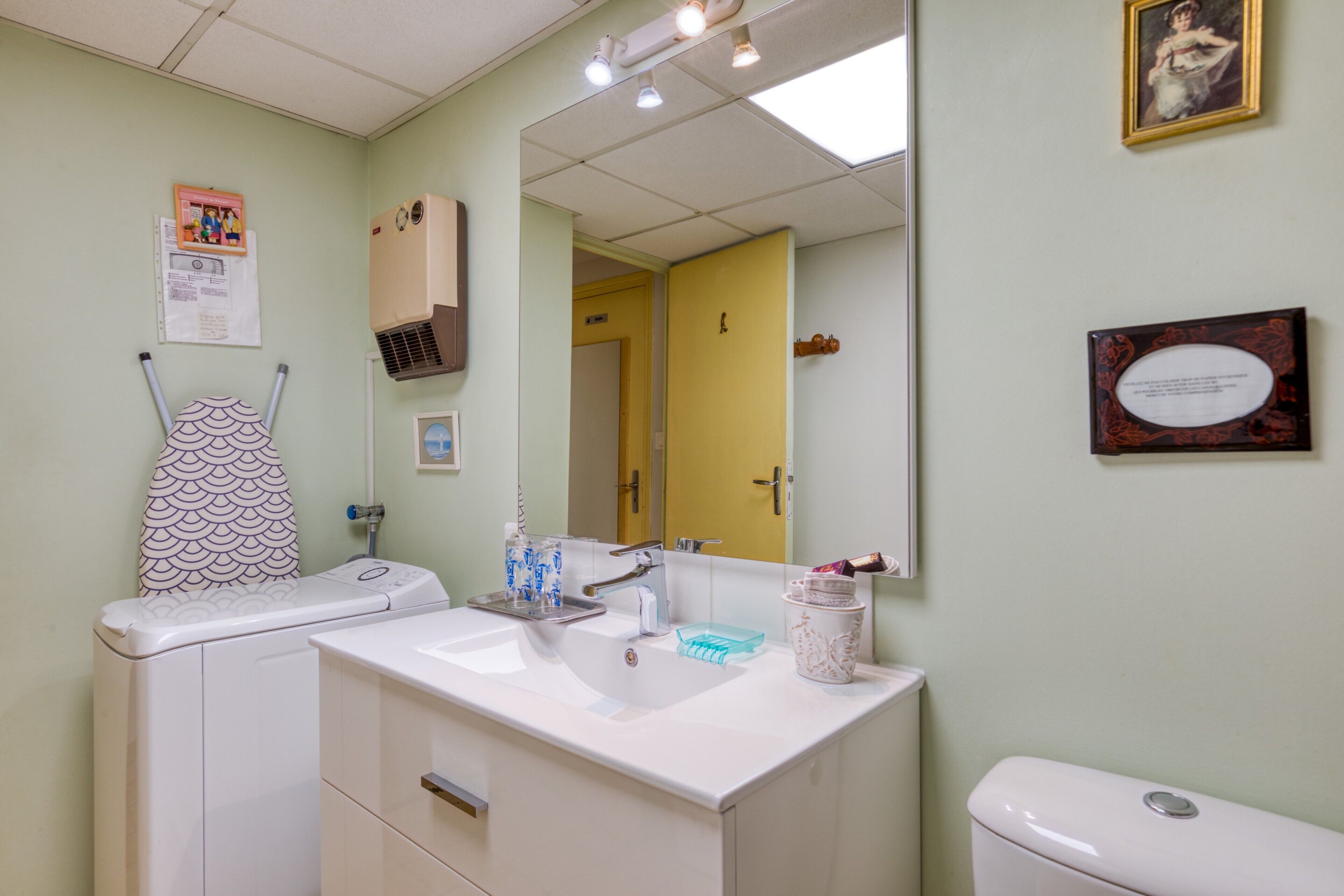 A bathroom with a sink, mirror, and toilet. There is a framed picture on the wall and a door leading to another room.