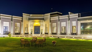 Un edificio moderno con entrada central, amplias ventanas y un césped bien iluminado con mobiliario exterior.