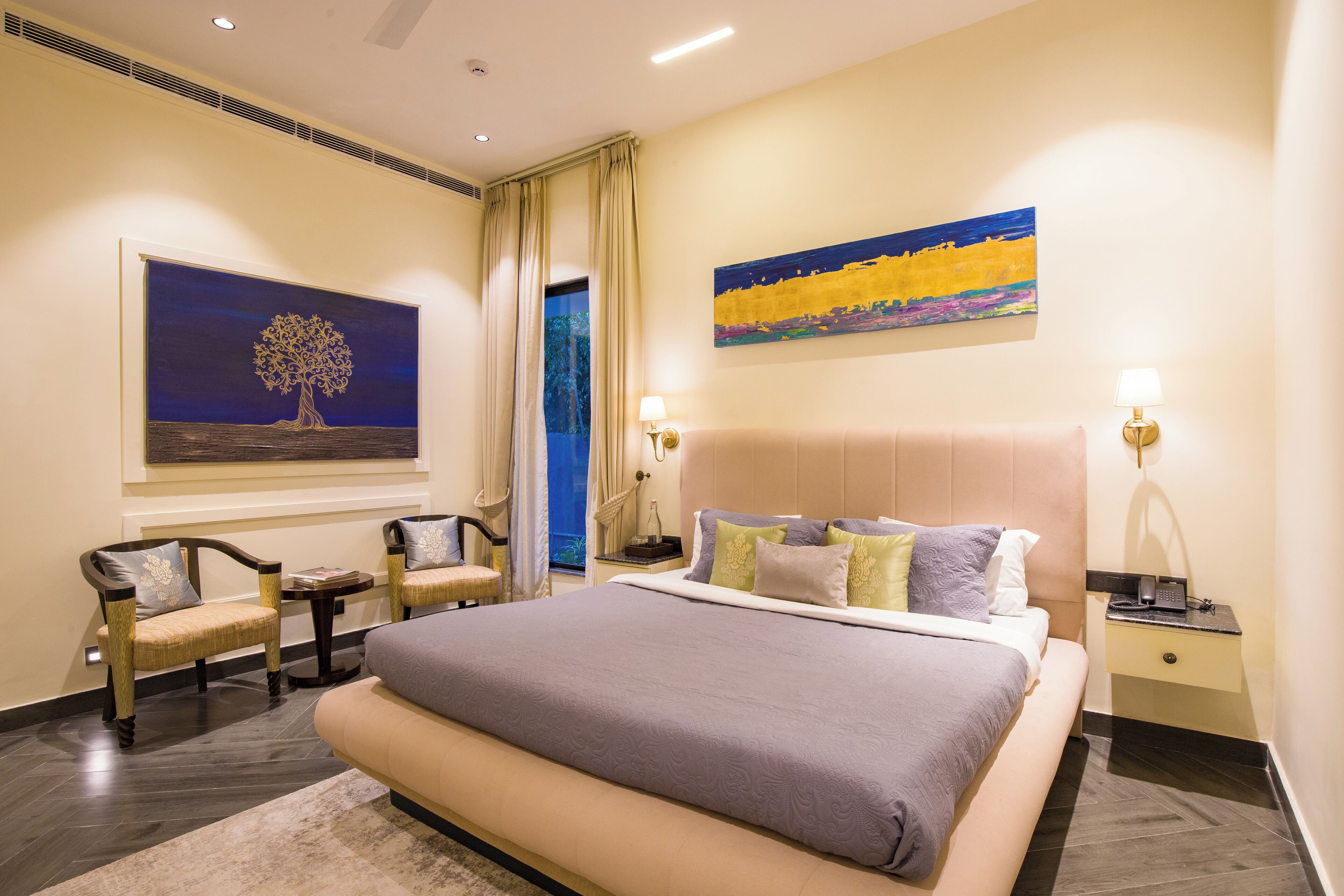 A hotel room with a large bed, two armchairs, a nightstand with a lamp, and artwork on the walls.