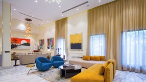 Un salón espacioso con un sofá amarillo, un sillón azul y un sofá beige. Se observa un cuadro grande en la pared, una araña y un ventanal con cortinas.
