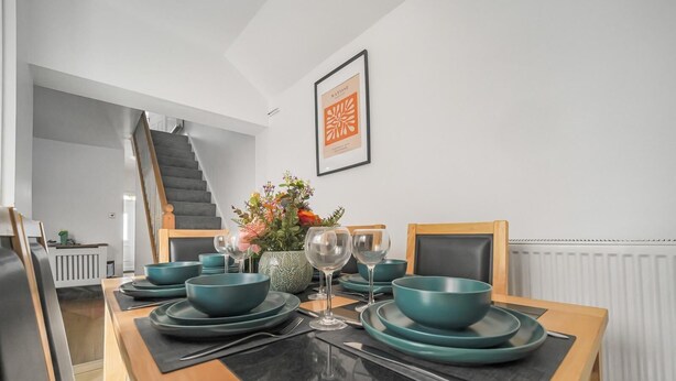 A dining area with teal dinnerware, a vase with flowers, and a framed artwork on the wall. - The Modern Cwtch House - Swansea (Gorseinon)