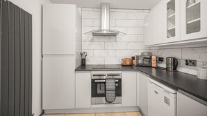 A modern kitchen with white cabinetry, a stainless steel range hood, a built-in oven, a dishwasher, and a countertop with a toaster, kettle, and coffee maker. - The Modern Cwtch House - Swansea (Gorseinon)