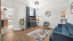 A modern living room with a blue sofa, a grey armchair, a wooden floor, a grey rug, a potted plant, a TV stand with a flat-screen TV, and a ceiling light fixture. - The Modern Cwtch House - Swansea (Gorseinon)