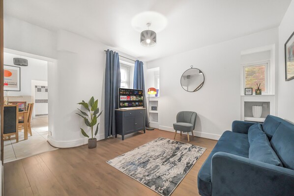 Una sala de estar moderna con un sofá azul, un sillón gris, suelo de madera, alfombra gris, una planta en maceta, un mueble de televisión con televisor de pantalla plana y una lámpara de techo.
