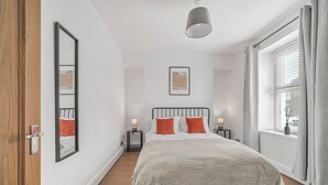 A bedroom with a bed, bedside tables, a mirror, a framed picture, and a window with curtains. - The Modern Cwtch House - Swansea (Gorseinon)