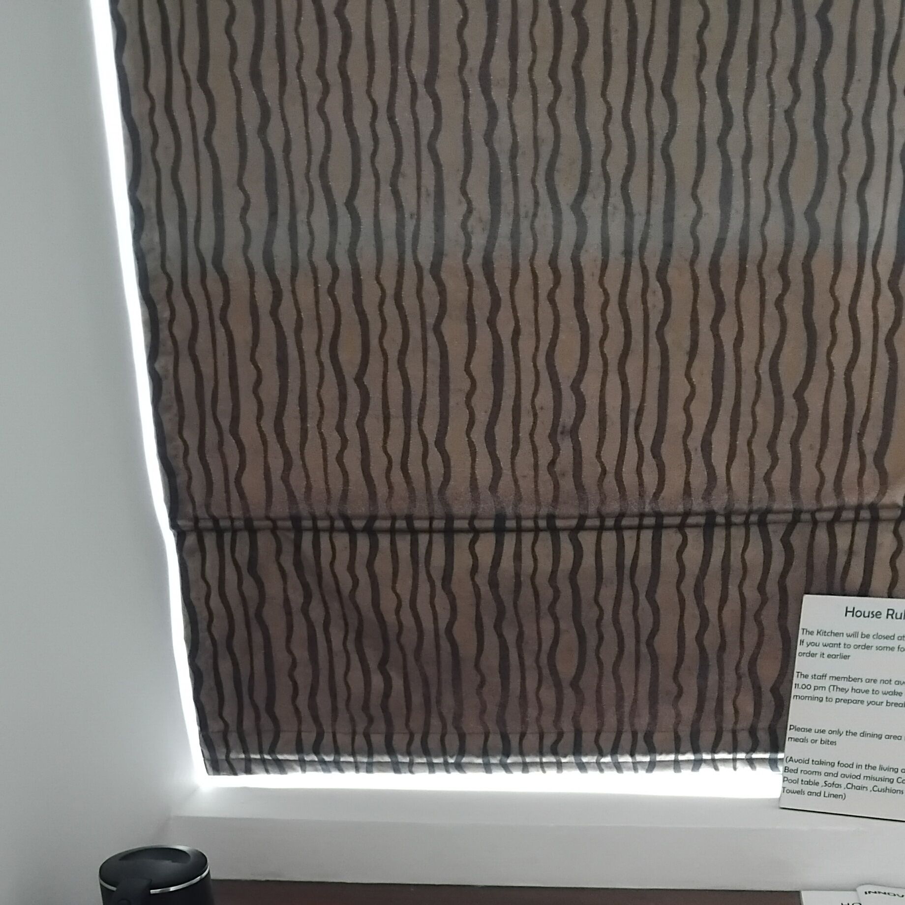 Una ventana con una persiana de diseño ondulado que la cubre parcialmente. Se ve un cartel con normas de la casa en el alféizar.