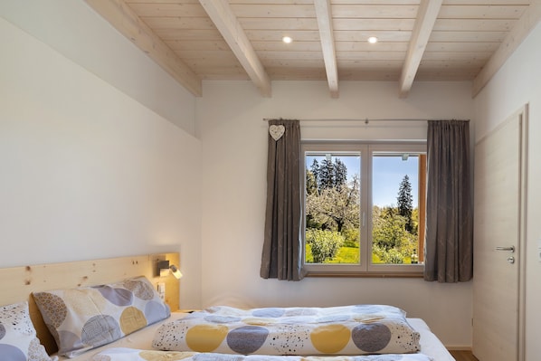 Un dormitorio con techo de madera, una cama con ropa de cama estampada, una ventana con cortinas y vistas a árboles.