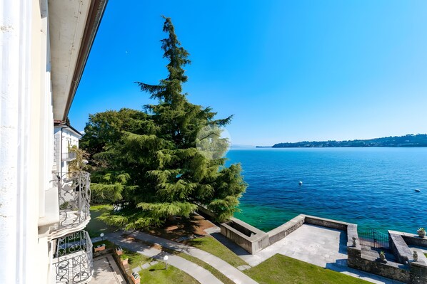 A balcony with a view of a clear blue lake, a large tree, and a stone pathway leading to a dock.