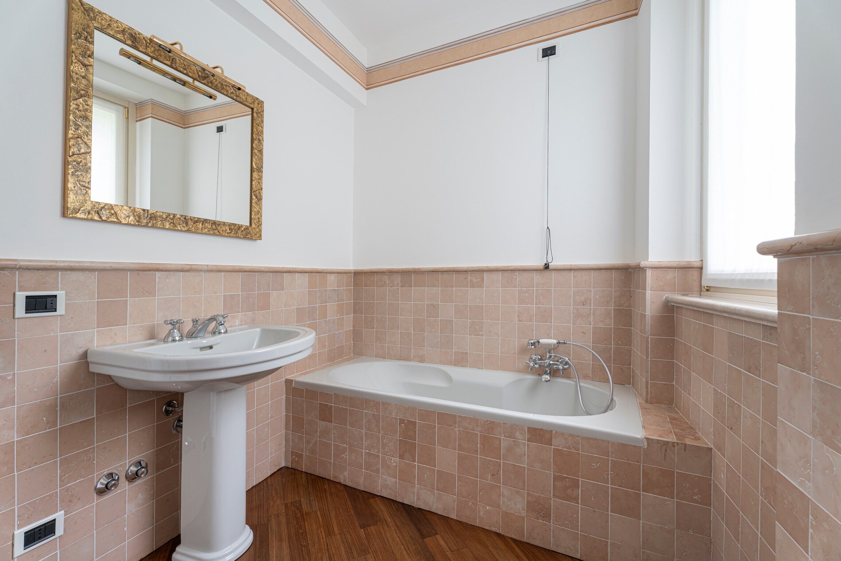 A bathroom with a pedestal sink, a bathtub, and a large mirror with a gold frame.
