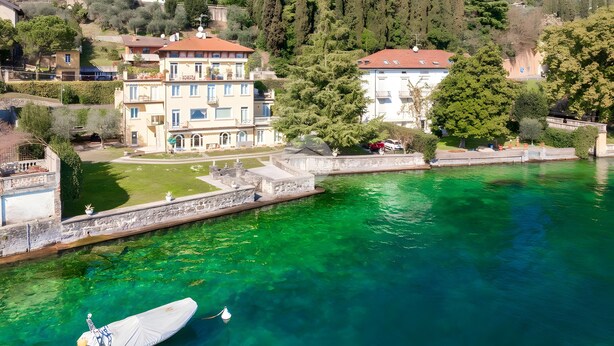 A waterfront property with a boat dock, a large building with multiple balconies, and a lush green hillside in the background. - Apartment 'La Gioconda' with Lake View, Wi-Fi and Air Conditioning (Salò)