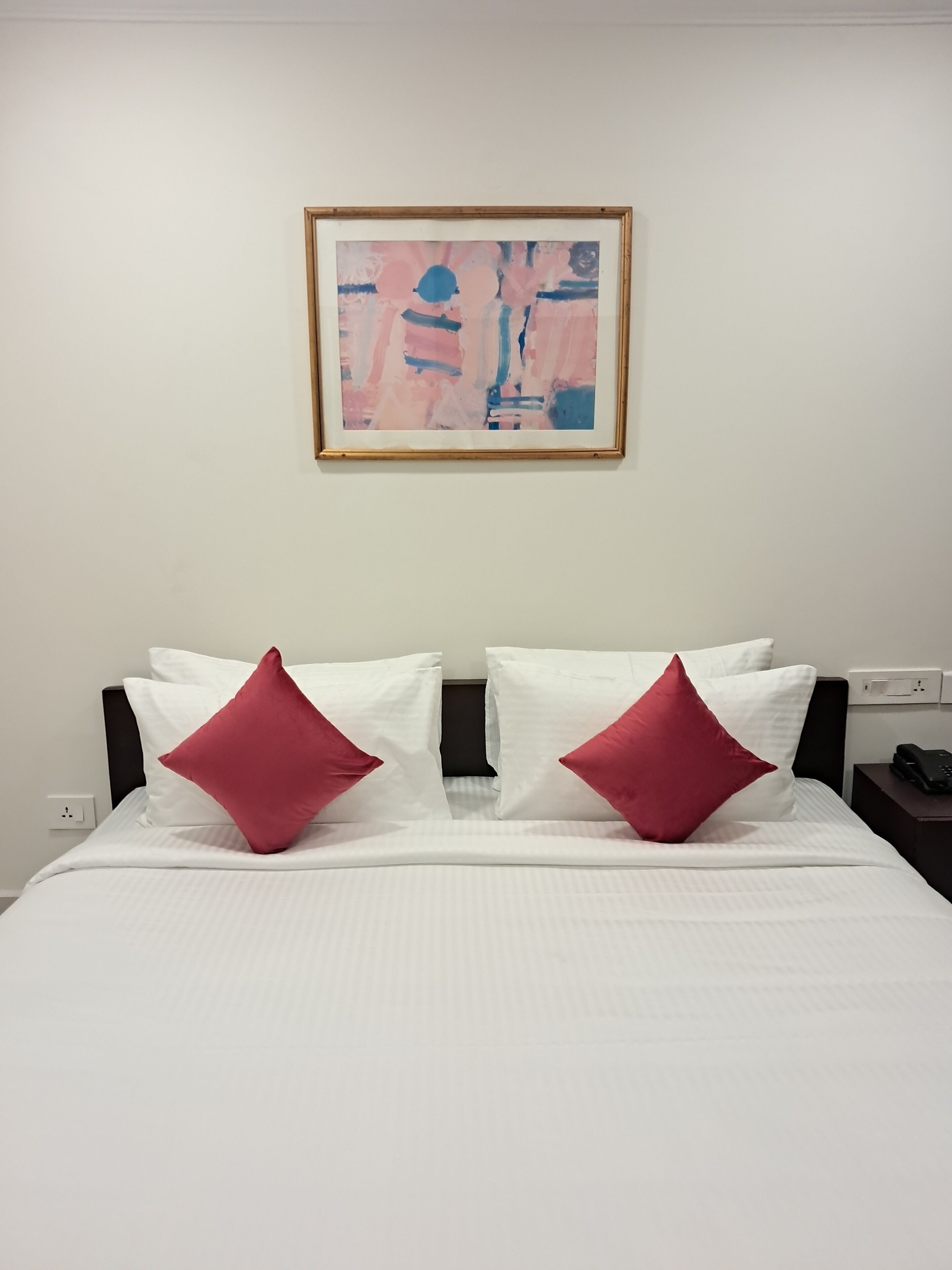 A neatly made bed with white bedding and two red pillows, a framed abstract painting on the wall, and a bedside table with a phone and charger.
