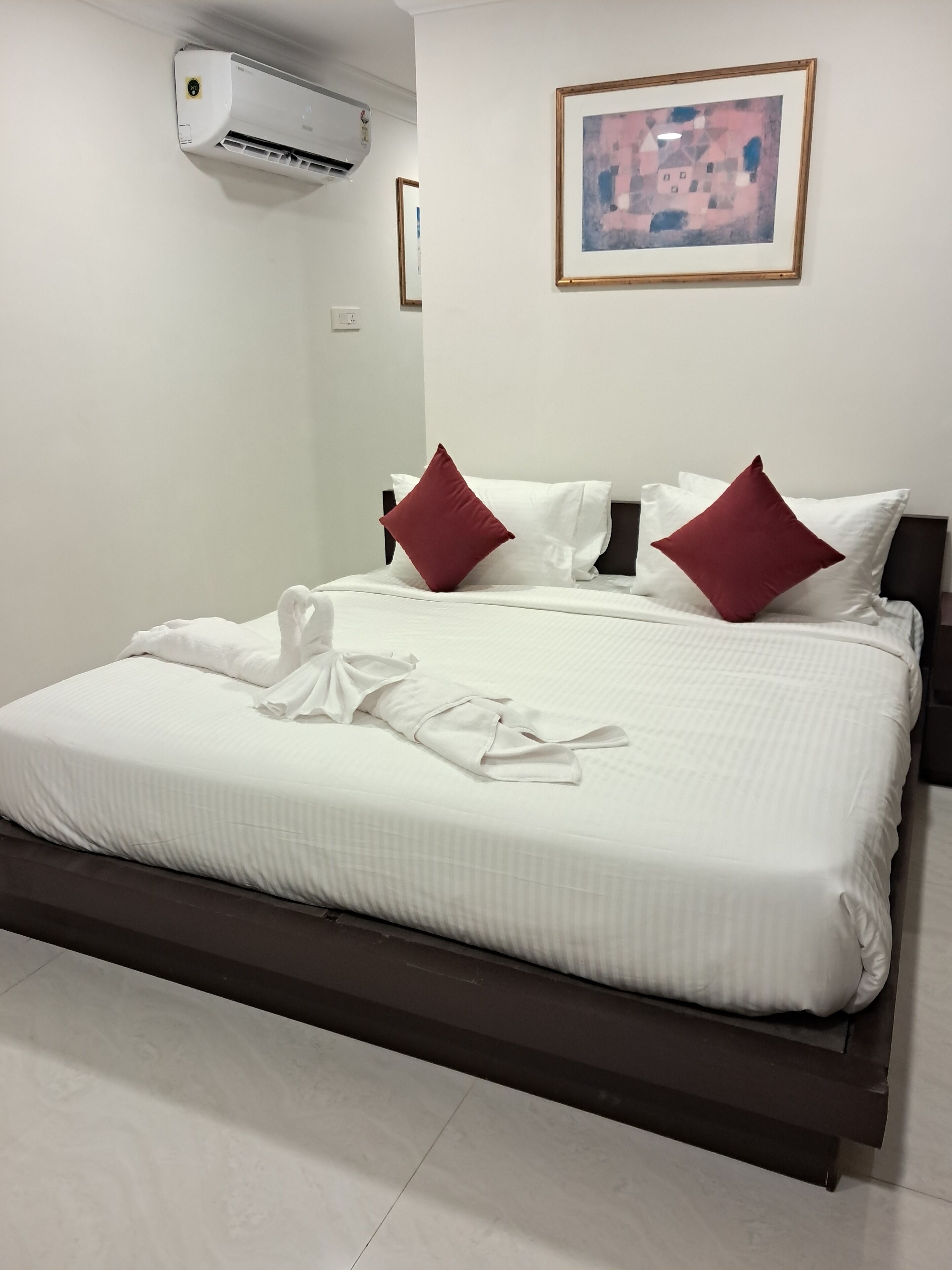 A neatly made bed with white linens and two red pillows, a framed abstract painting on the wall, and a white air conditioning unit above the bed.