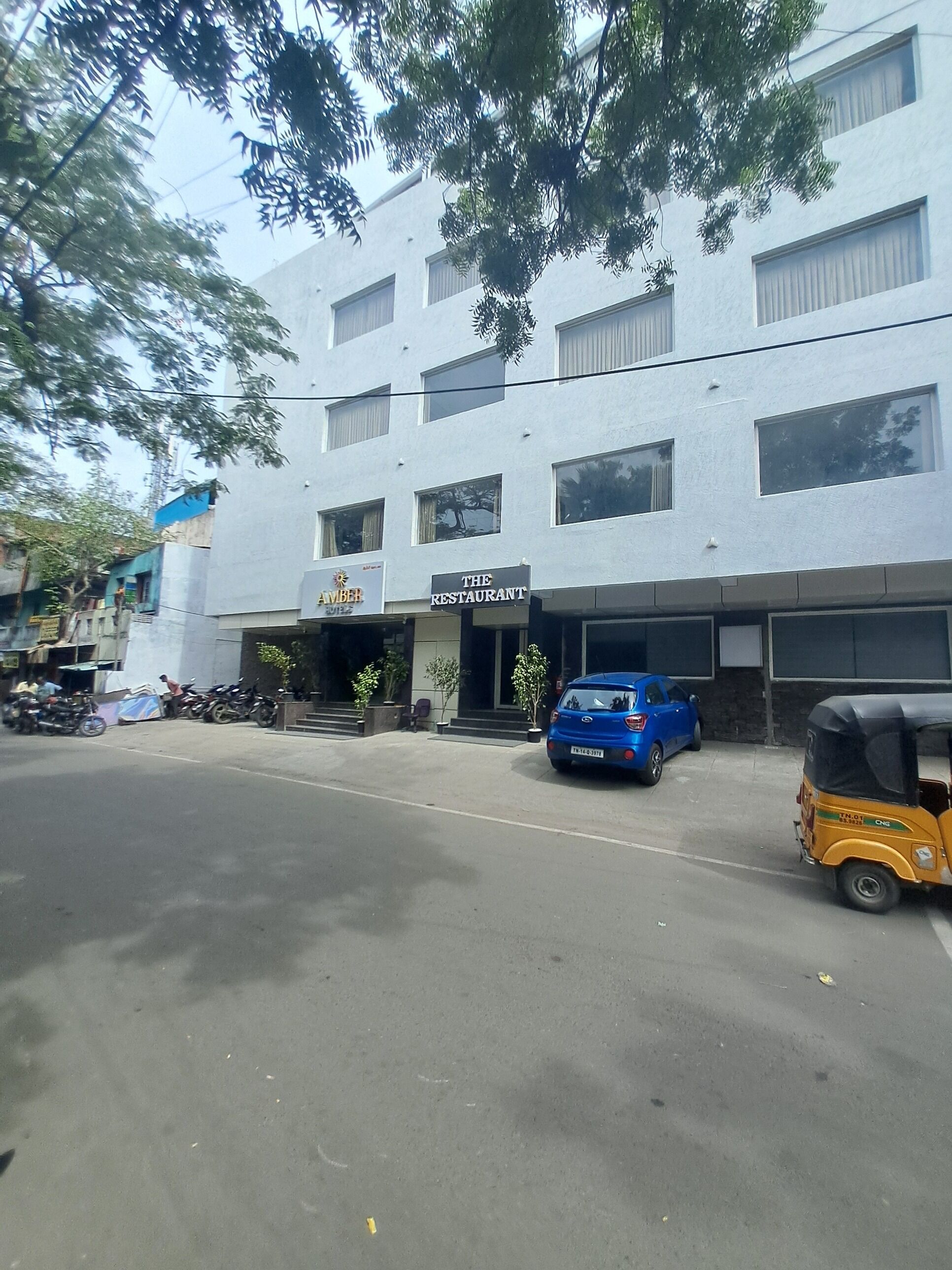 A building with a sign for "The Restaurant" and "Amber" on the ground floor. There are parked vehicles and a yellow auto-rickshaw in front.