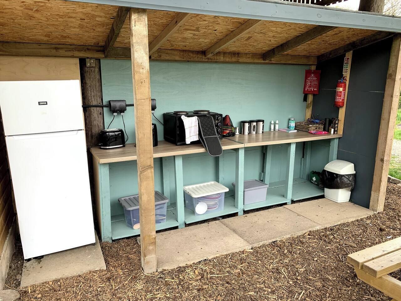 A small outdoor kitchen area with a refrigerator, a coffee maker, and various containers.