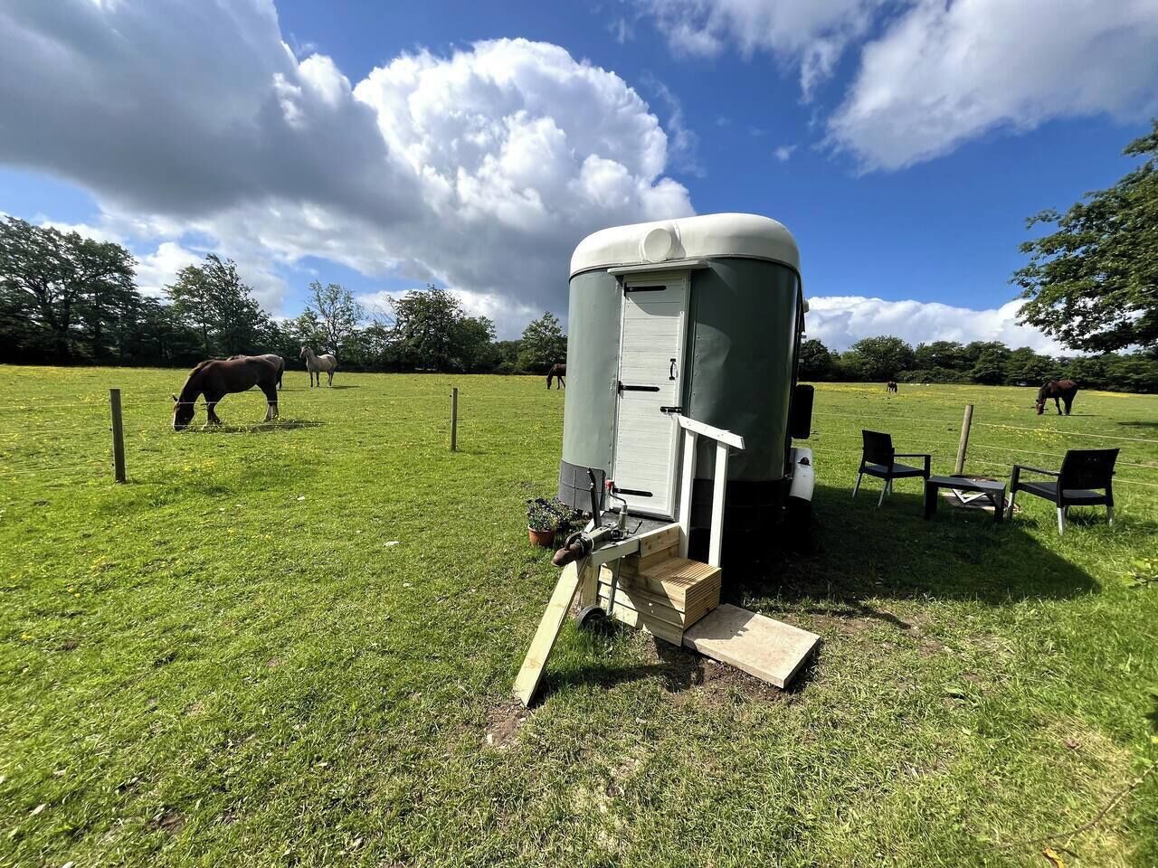 Des toilettes portables installées dans un champ herbeux, avec quelques chaises et une structure en bois à proximité.