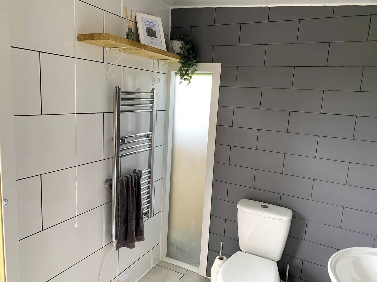 Une salle de bain avec des murs carrelés de couleur grise, des toilettes blanches, un porte-serviettes et un espace douche.