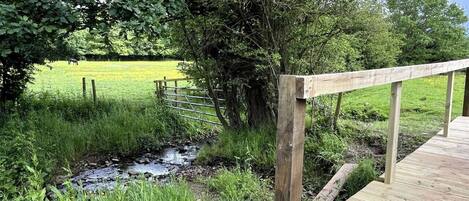 A wooden deck with a metal railing overlooking a grassy field and a small stream.
