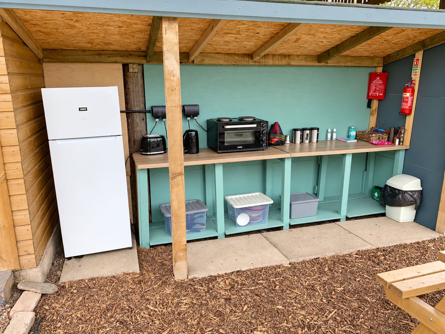 Una cocina al aire libre compacta con refrigerador, microondas, tostadora y cafetera. Hay contenedores de almacenamiento debajo del mostrador y un extintor montado en la pared.