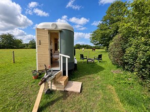 Une maison mobile, dont la porte est ouverte, dévoile une chambre, un petit espace extérieur aménagé pour s’asseoir, et un champ verdoyant avec des arbres en arrière-plan.