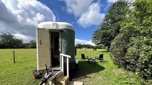 Une petite maison mobile d’un seul étage, avec une porte ouverte, une terrasse en bois et quelques plantes en pot.