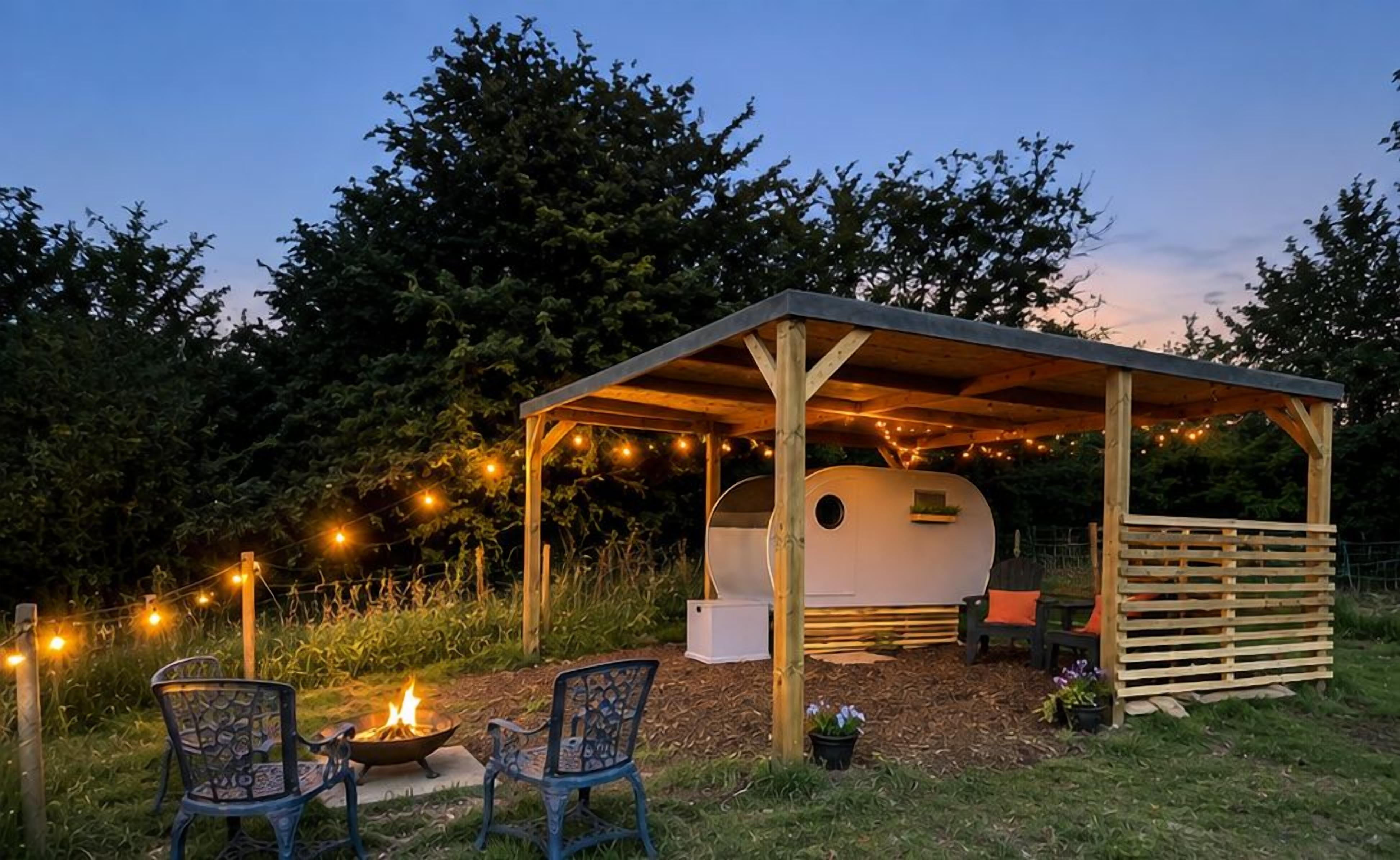 Un área de estar al aire libre cubierta con una chimenea, sillas y una caravana bajo una pérgola de madera con luces colgantes.