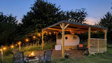 Un área de estar al aire libre cubierta con una chimenea, sillas y una caravana bajo una pérgola de madera con luces colgantes.