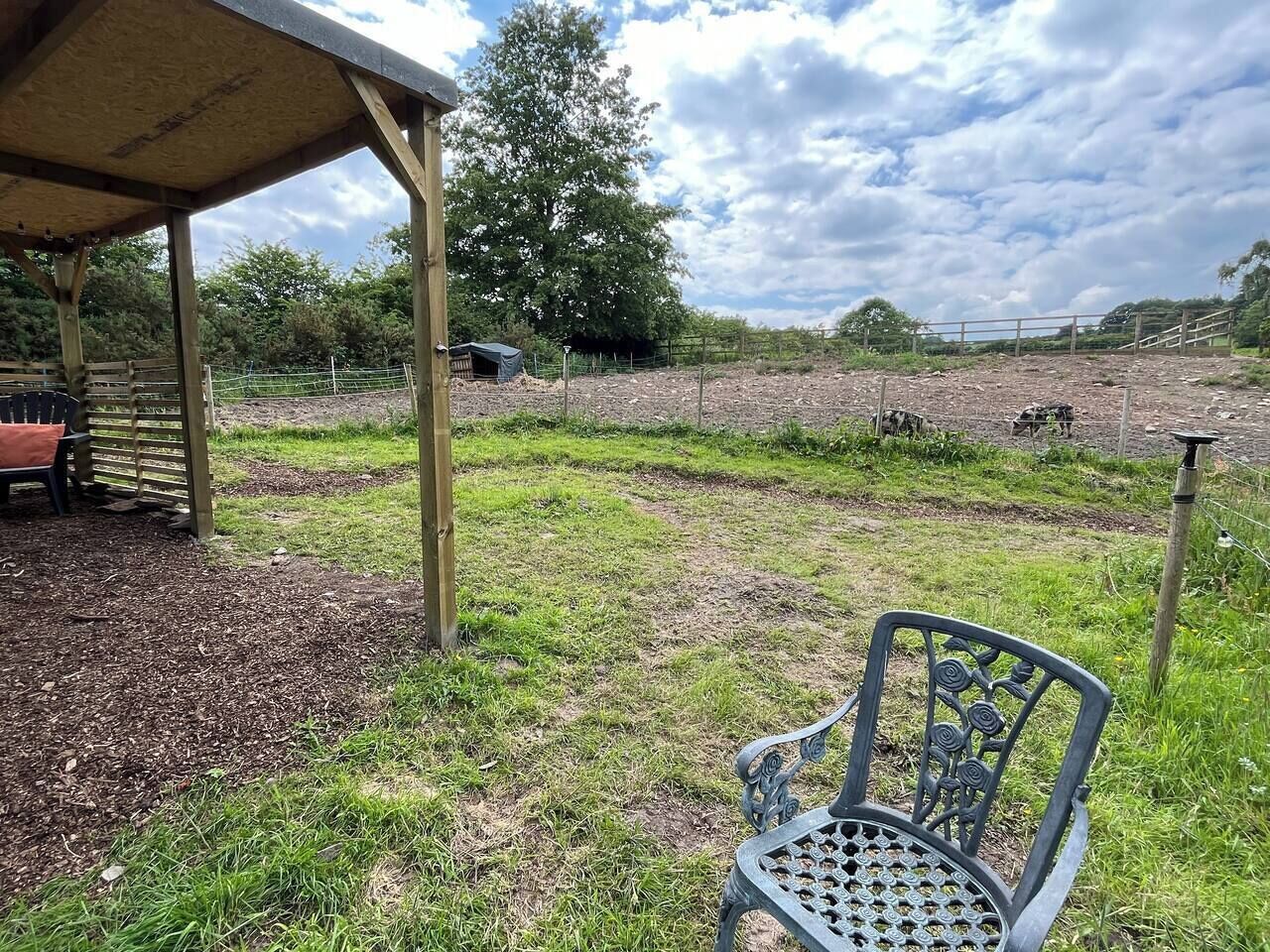 A covered outdoor area with a metal chair, a wooden bench, and a fenced field.