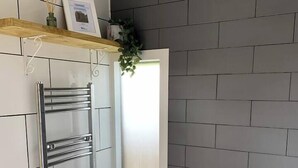 A bathroom with a grey tiled wall, a white toilet, a towel rack with a towel, and a small shelf with a plant and a picture. - The Pig Sty - Farm Stay (Shepshed)
