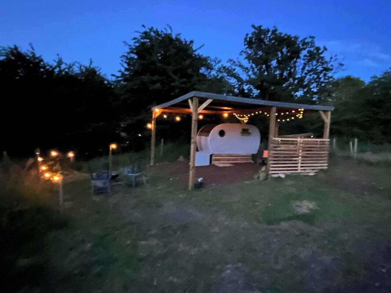 A rustic outdoor structure with a wooden roof and string lights, surrounded by grass and trees.