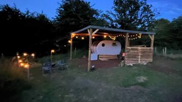 A rustic outdoor structure with a wooden roof and string lights, surrounded by grass and trees.