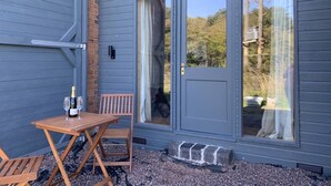 A wooden table with two glasses and a bottle, a bench, and a sliding glass door with a view of a garden. - The Barn - Farm Stay (Shepshed)
