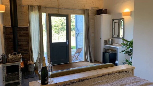 A cozy room with a bed, a wooden bench, a fireplace, a door leading outside, and a small table with a bottle and glasses. - The Barn - Farm Stay (Shepshed)
