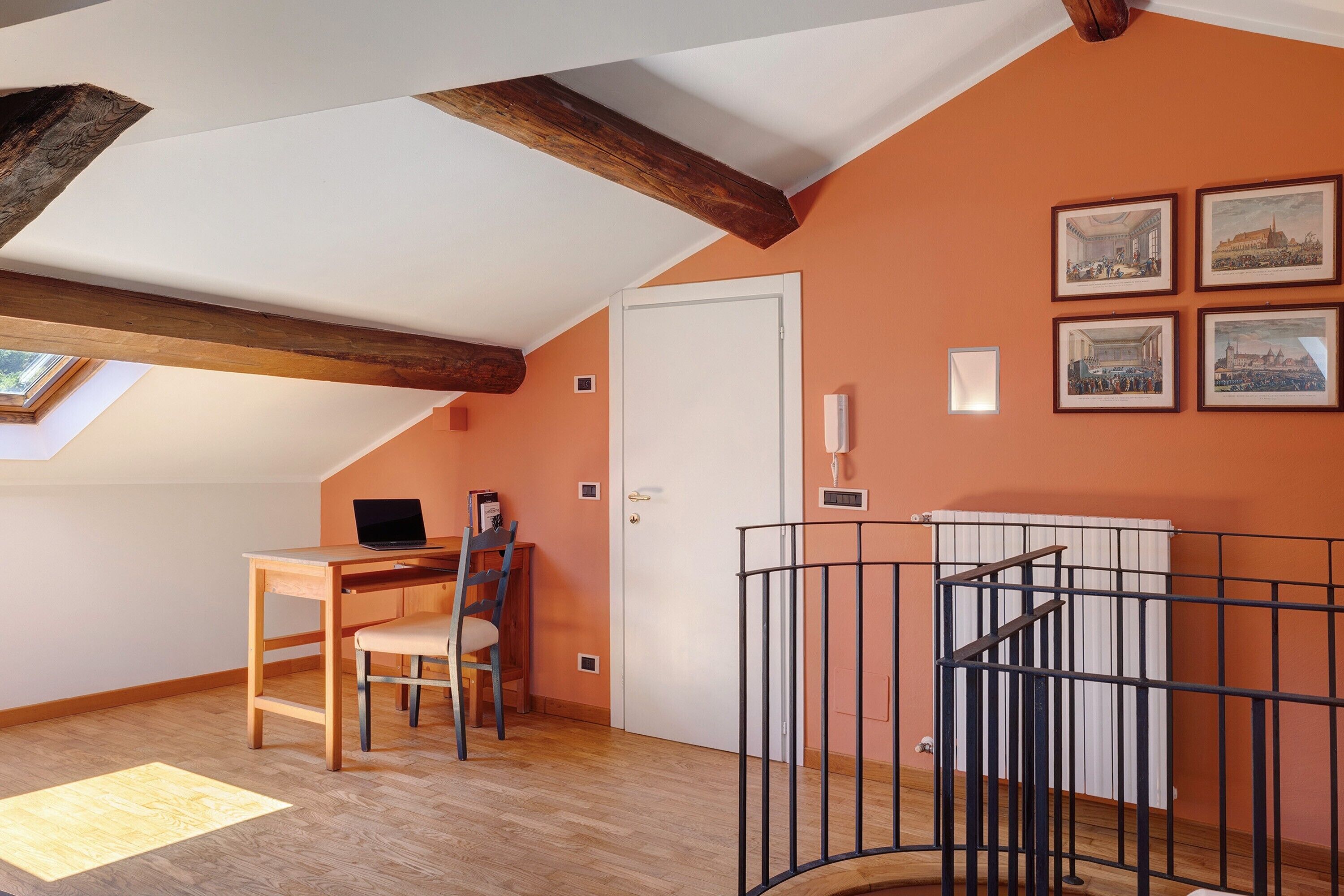 A room with a sloped ceiling, wooden beams, a desk with a chair, a laptop, and framed pictures on the wall.