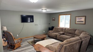 Ein Wohnzimmer mit Ecksofa, einem Holzschaukelstuhl, einem kleinen Tisch, einem an der Wand montierten Fernseher und einem Fenster mit Jalousien.