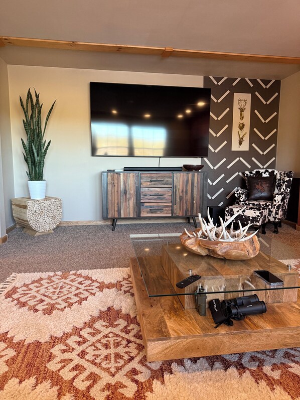 A modern living room with a large flat-screen TV, a wooden coffee table with antlers, a patterned rug, and a potted plant.