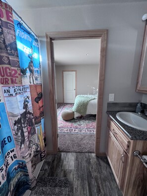 A bathroom with a wooden vanity, a sink, and a mirror. There is a door leading to a bedroom with a bed and a window.