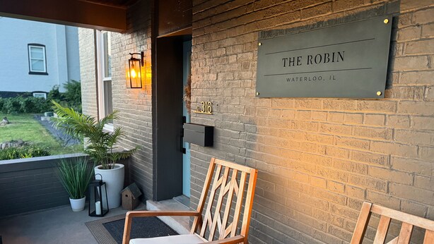 Ein hölzerner Schaukelstuhl mit Kissen auf einer Veranda mit einer Ziegelwand und einem Schild, auf dem 'The Robin Waterloo, IL' steht.