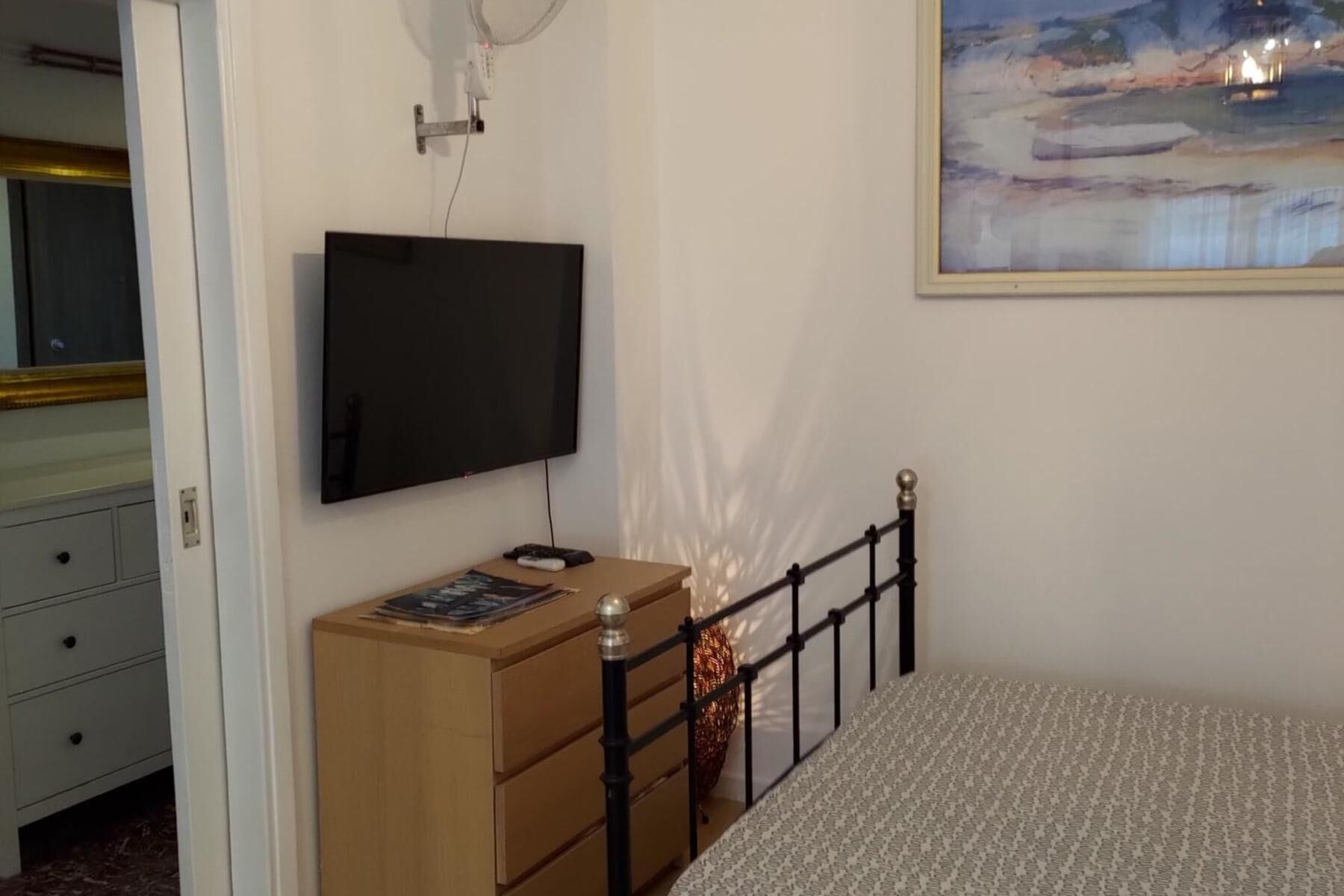 A bedroom with a bed, a dresser, a television mounted on the wall, and a painting above the bed.