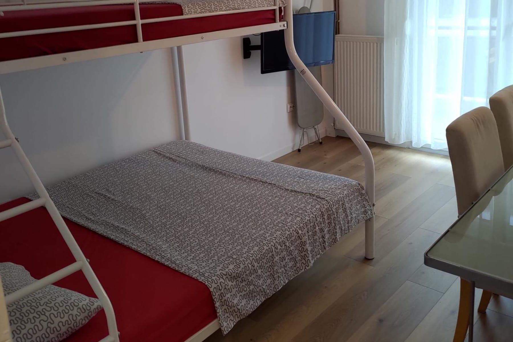 A bunk bed with a red mattress and a patterned blanket, a small table, and a chair in a room with wooden flooring and a window with curtains.