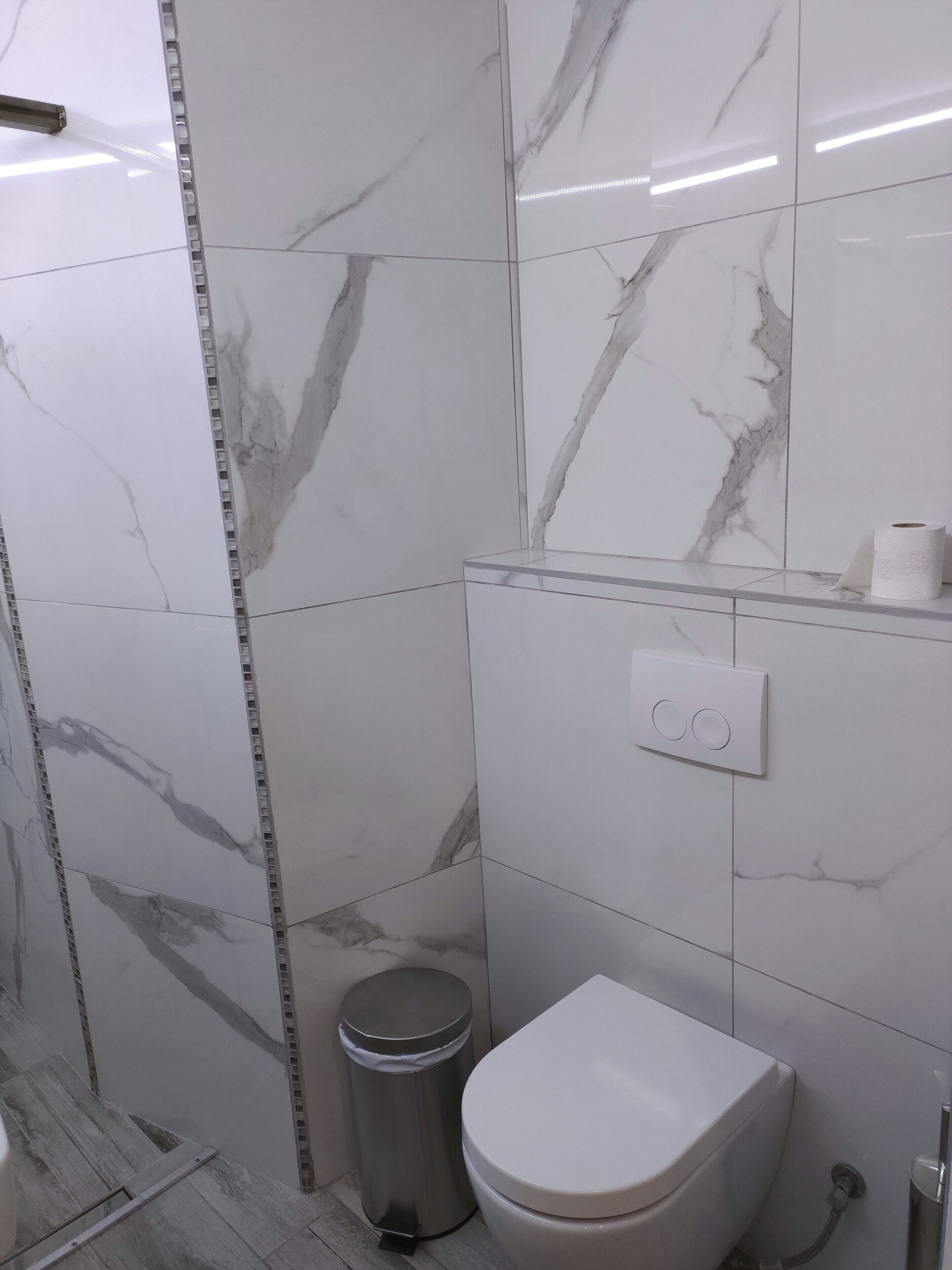 A bathroom with white marble tiles, a toilet, a trash bin, and a light switch.