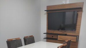 Een eetgedeelte met een witte tafel en vier stoelen, een houten tv-meubel met een flatscreen televisie en een betegelde vloer.