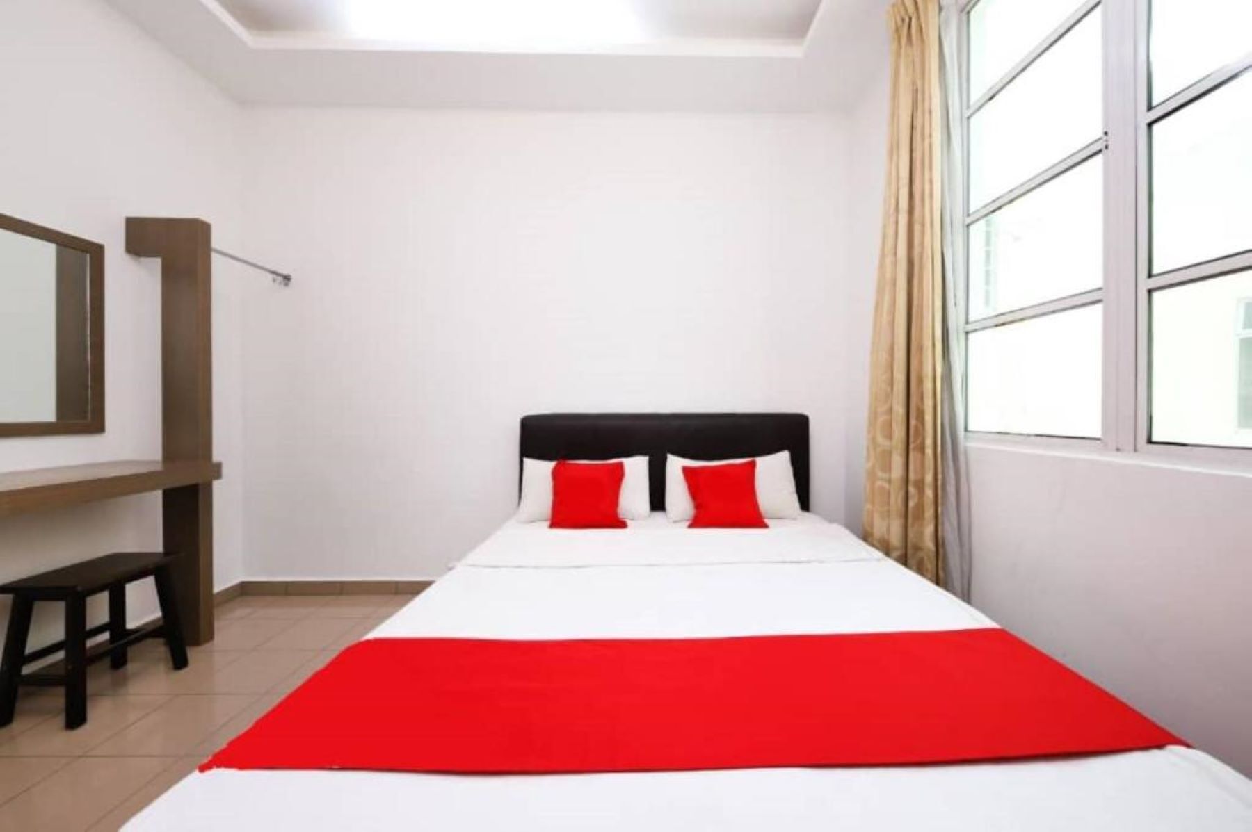 Un lit simple avec une couvre-lit rouge et blanche, deux oreillers rouges, une tête de lit noire, un banc en bois et une grande fenêtre avec des rideaux.