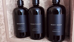 Three black bottles of shampoo, conditioner, and body wash in a marble-tiled bathroom.