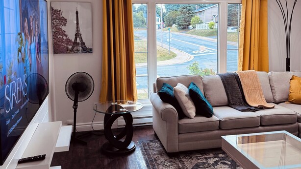 A modern living room with a large flat-screen TV, a sofa with cushions, a coffee table, a standing fan, and a large window with curtains.