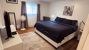 A bedroom with a bed, a desk, a chair, a standing fan, a window with curtains, and a ceiling light.