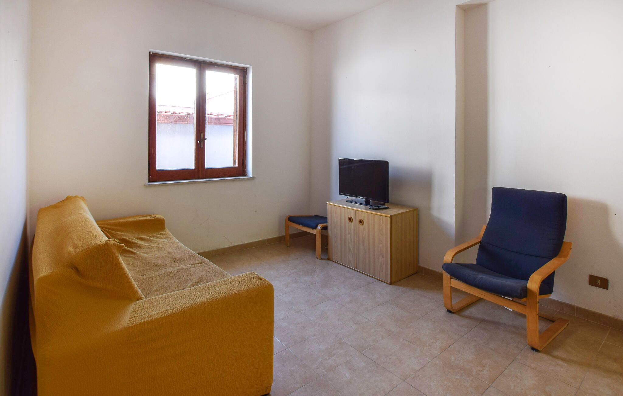Uma sala de estar com sofá, televisão sobre um armário de madeira, uma poltrona e janela com vista para prédios.